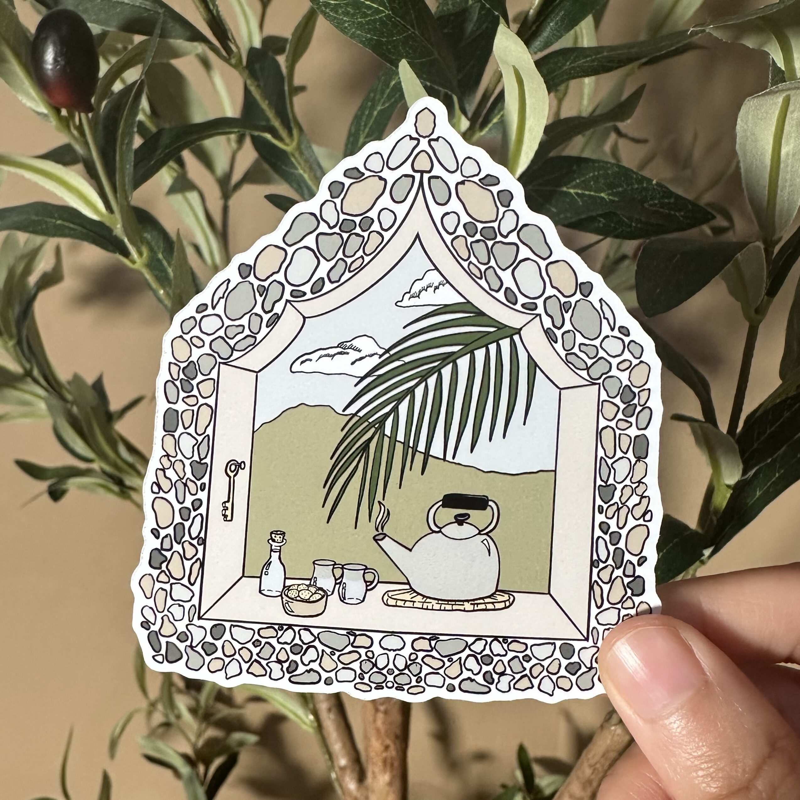 a person holding a 2x3" sticker with the olive tree as a background